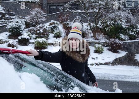 Young attractive brunette woman clearing snow off a car with a ice scraper in the winter. Stock Photo