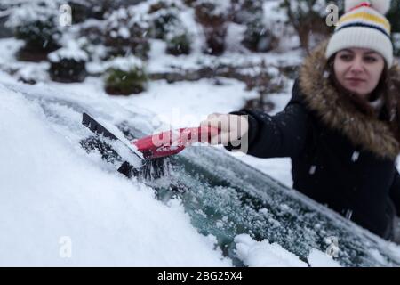 Young attractive brunette woman clearing snow off a car with a ice scraper in the winter. Stock Photo