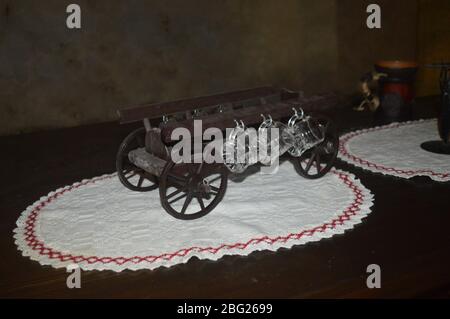 A rustic miniature wooden wagon used as a novelty cup holder for small glass mugs, displayed on a traditional embroidered doily atop a wooden table. Stock Photo
