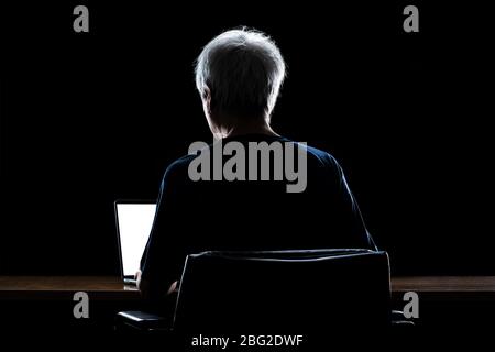 Back view of a man with gray hair working from home late at night using his laptop computer, back lit, work from home concept Stock Photo