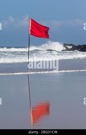 A red Safety flag flapping in the wind with large waves breaking on the rocks behind. Stock Photo