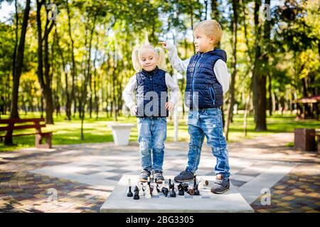 Funny little brother and sister of Caucasian ethnicity play on a public chess venue in a city park. Children frolic and indulge instead of studying Stock Photo