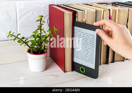 Woman hand takes e-reader from shelf with paper books and small potted plant. Copy space on e-book display. E-reading for pleasure and education. Stock Photo
