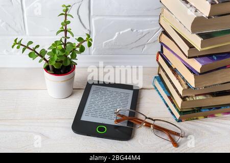 E-book reader and vision glasses near stack of paper books and small potted plant on a white wooden table. E-reading for pleasure and education. Stock Photo