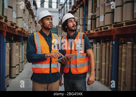 Young sacred workers in warehouse discussing work on digital tablet looking away towards goods on shelf Stock Photo