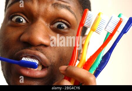 A young man brushes his teeth with one hand, whilst holding a number of toothbrushes in his other hand. Stock Photo
