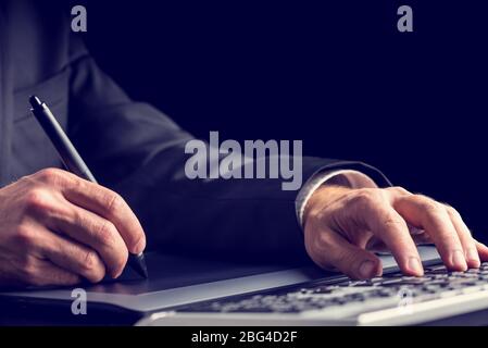 Businessman typing on a computer keyboard while simultaneously using a tablet and stylus to navigate or be creative, closeup view of his hands. Stock Photo