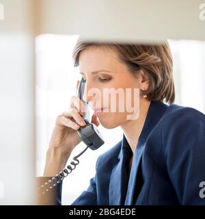 View through an internal office partition of a successful young businesswoman sitting at her desk making a phone call on a landline telephone, profile Stock Photo