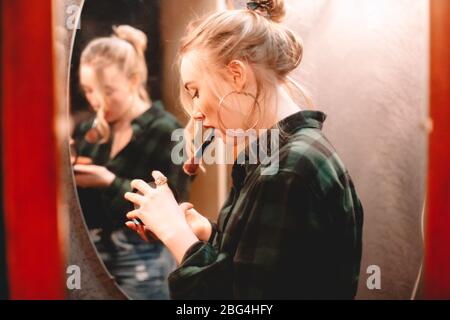 Young woman opening palette and holding makeup brush in her mouth while applying make up standing in front of mirror at home getting ready to go out
