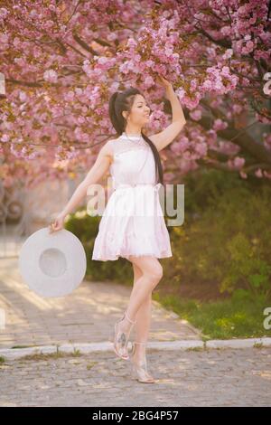 Young woman in white dress and hat walks and enjoys in park at alley with blooming sakura trees. Stock Photo