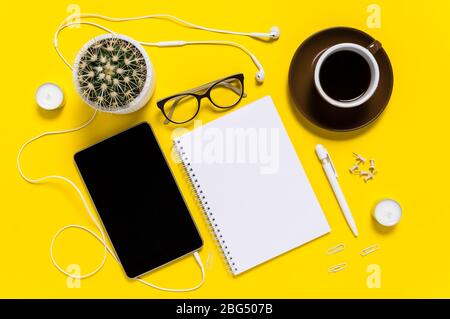 Flat lay of modern home office workspace with tablet, coffee cup, note pad, pen, eyeglasses and cactus on yellow background. Top view Stock Photo