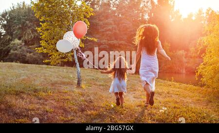 Mother's day. Little girl running with mother and holding baloons in hand. Family having fun in summer park Stock Photo