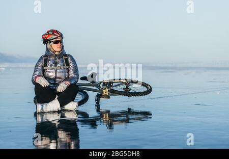 Woman sitting near her bike on ice. The girl cyclist stopped to rest. She sits on the wheel and enjoys the beautiful view of the sunset. Stock Photo