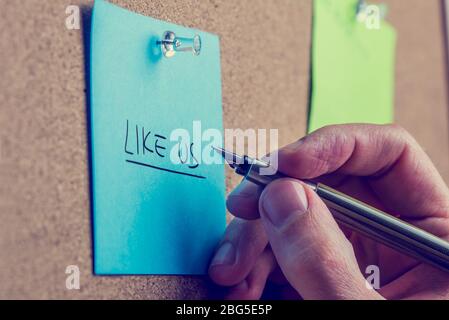 Man writing - Like Us - on a blue note notice board in a request for followers on social media sites and networks. Stock Photo