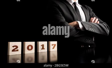 Cropped upper body of businessman in dark suit, tie and white shirt with folded arms and year 2017 on blocks. Stock Photo