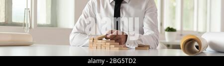 Businessman seated at a table building steps of wooden blocks in a wide panorama view of his hands across the surface with copy space. Stock Photo