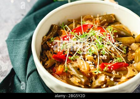 Stir fry glass rice noodles with vegetables, green beans, carrot, bell peppers, sesame seeds and microgreen in bowl on gray background. Close up. Stock Photo