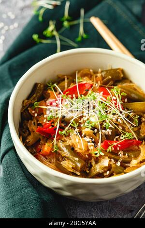 Stir fry glass rice noodles with vegetables, green beans, carrot, bell peppers, sesame seeds and microgreen in bowl on gray background. Close up. Stock Photo