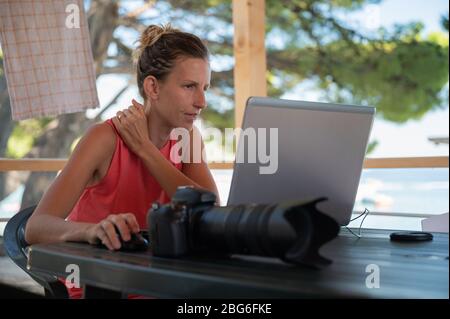 Young female photographer editing phots on her laptop computer while on vacation in summer beach resort. Stock Photo