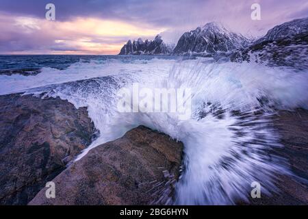 Huge waves crashing onto the coastal rocks at high tide in the morning, with colorful sky and jagged snow mountains on the horizon.