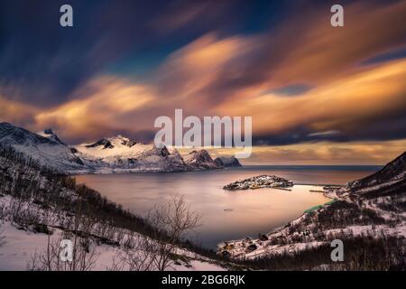The warm sunset light casting on clouds, streaking over the snowcapped mountains and beautiful fjord, with an fishing village on an island. Stock Photo