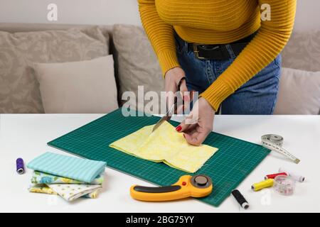 woman in a yellow cuts the fabric. there are quilting tools on the table. patchwork knife, scissors, lined cutting mat, self-locking, thread, centimet Stock Photo