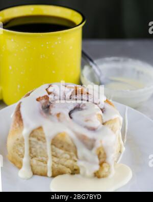 Cinnamon roll on a white plate with cup of coffee and bowl of glaze in the background. Stock Photo