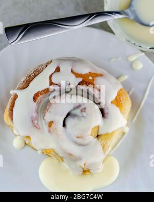 Overhead shot of a cinnamon roll on a white plate with a bowl of glaze. Stock Photo