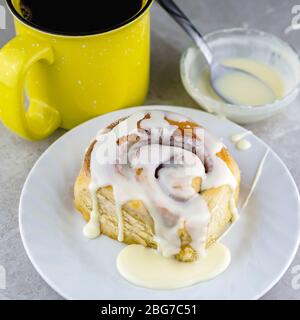 Cinnamon roll on a white plate with cup of coffee and bowl of glaze in the background. Stock Photo
