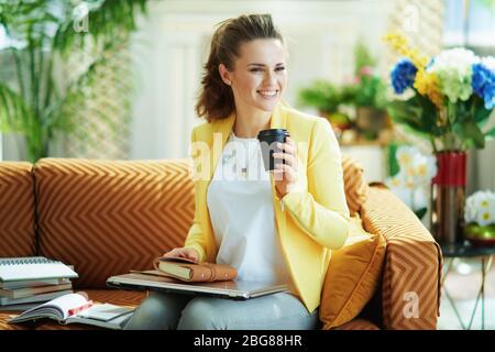 smiling young learner woman in jeans and yellow jacket sitting on couch with a closed laptop and coffee ready to study online at modern home in sunny Stock Photo