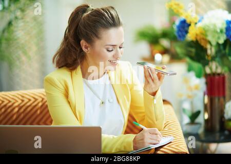 smiling stylish housewife in jeans and yellow jacket with laptop and smartphone study online at modern home in sunny day. Stock Photo