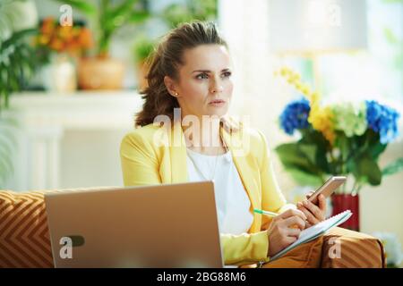 pensive elegant 40 years old woman in jeans and yellow jacket with laptop and smartphone study online in the modern living room in sunny day. Stock Photo