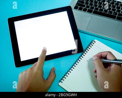 Hand pointing on white blank space screen on tablet and hand writing on notebook with laptop computer on blue background, top view. Stock Photo