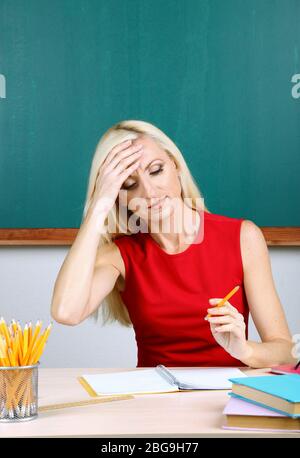 Tired teacher sitting at table on blackboard background Stock Photo