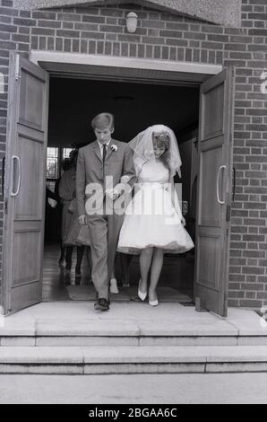 1960s, historical, a 'teenage' bride in her wedding dress with bridal veil and her groom in a three button suit of the era, walking out of the modern church building following their wedding, England, UK. Stock Photo