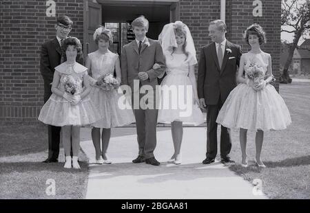 1960s, historical, group picture showing wedding guests with a 'teenage' bride in her wedding dress with bridal veil and groom in a three button suit of the era, standing together outside the entrance to a modern church building, England, UK. Stock Photo