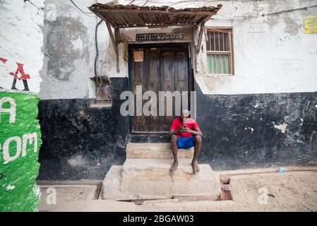 Lamu, Kenya - December 15, 2016: young black African man using his phone sitting in front of door in Lamu town Stock Photo