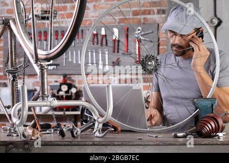 man repair the vintage bicycle with mobile phone at the computer search spare parts on the internet in garage workshop on the workbench with tools, di Stock Photo