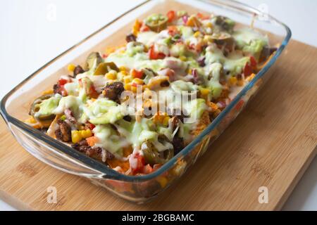 Baked nachos with corn tortilla chips, corn, guacamole, pico de gallo, beans, jalapenos and Monterrey jack in a glass baking dish Stock Photo