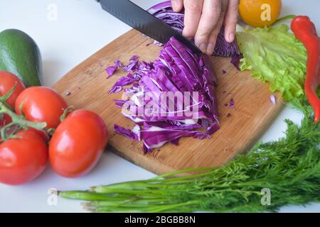 A man cutting red cabbage in ribbons on a wooden cutting board surrounded with tomatoes, avocado, lettuce, dill, lemon and red jalapeno pepper Stock Photo