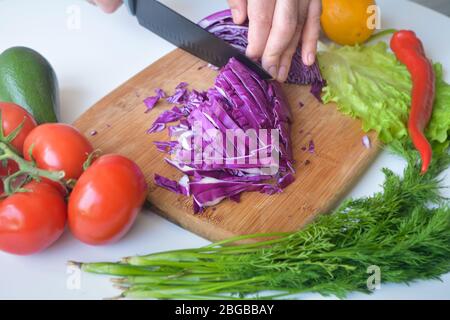 A man cutting red cabbage in ribbons on a wooden cutting board surrounded with tomatoes, avocado, lettuce, dill, lemon and red jalapeno pepper Stock Photo