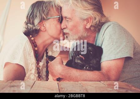 Happy old senior couple people in love kiss eachother and hug their lovely black dog pug - home leisure activity and forever together concept family a Stock Photo