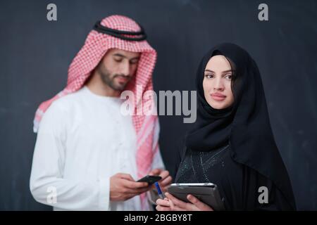 young muslim business couple arabian man with woman in fashionable hijab dress using mobile phone and tablet computer in front of black chalkboard rep Stock Photo