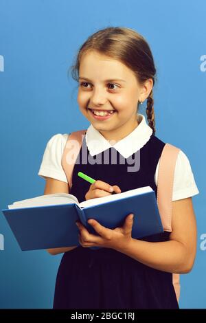 Back to school and education concept. School girl with happy smiling face isolated on blue background. Pupil in school uniform with braid and backpack. Girl writes in big blue notebook with pen Stock Photo