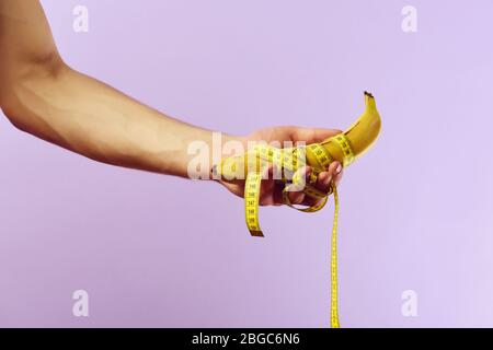 Fitness, regime and diet concept. Hand of man holding banana wrapped around with yellow measuring tape, on purple background. Weight management idea Stock Photo