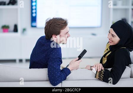 young muslim couple woman wearing islamic hijab clothes sitting on sofa watching TV together during the month of Ramadan at modern home Stock Photo