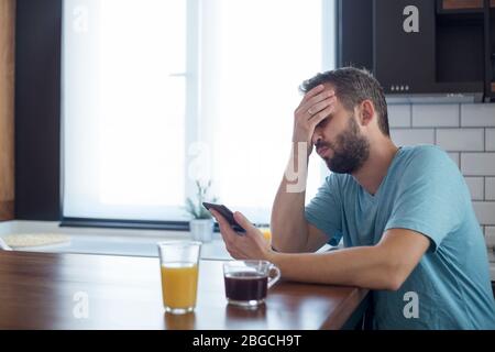 Young man disappointed while working on tablet. He is sitting at his kitchen table at home. Stock Photo