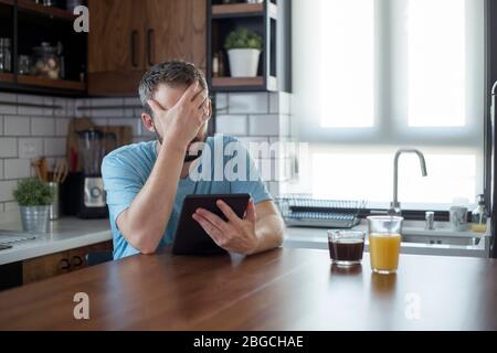 Young man disappointed while working on tablet. He is sitting at his kitchen table at home. Stock Photo