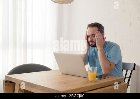 Young man disappointed while working on laptop. He is sitting at table at home. Stock Photo