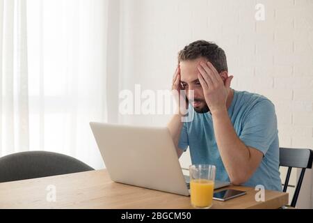 Young man disappointed while working on laptop. He is sitting at table at home. Stock Photo